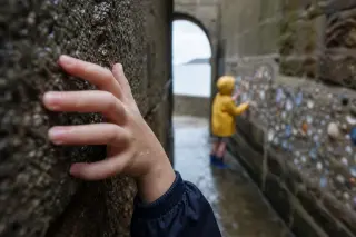 Child's hand on rain-damp granite wall under Saint-Malo ramparts, another child in yellow raincoat exploring shells in background, natural Breton light highlighting stone textures.