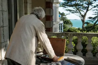 Elderly artist from behind arranging paints on a villa veranda in Saint-Briac-sur-Mer, with garden and sea in soft morning light