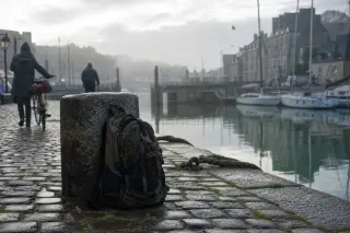Misty Breton quay at dawn with a travel backpack against a granite mooring post, wet cobbles and blurred locals passing by, soft silver morning light
