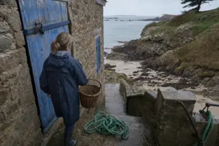 Worn granite steps by a stone house in Rothéneuf leading to a cove, with a woman in a raincoat holding a basket, early morning light revealing damp textures and sea spray.