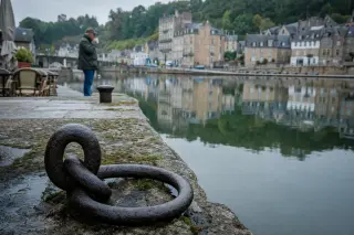 Wet granite mooring ring on old quay at Dinan port, with man drinking coffee on terrace and blurred reflections of historic buildings in calm river behind