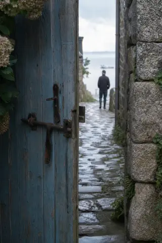 Weathered wooden gate ajar on a damp cobbled alley in Saint-Suliac with a fisherman walking toward the river, soft morning light highlighting moss, hydrangeas, and old stone walls.