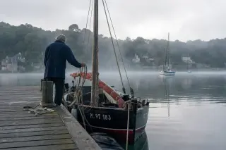 Mature sailor in navy pullover preparing a boat on a wooden pontoon at dawn on the misty Rance river, with soft morning light and distant village visible