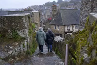Two locals in raincoats descend worn stone steps of Dinan's medieval ramparts after rain, with mossy granite walls and misty rooftops in the background.