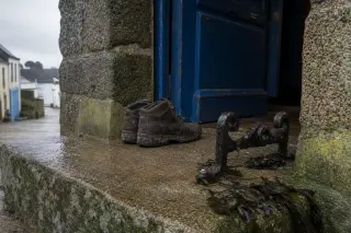 Granite doorstep with weathered boot scraper, muddy boots, seaweed, and a blue wooden door ajar in a Breton village, under soft morning light after rain.