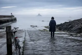 A woman in a raincoat stands on a wet granite slipway in a Breton port, gazing toward the misty silhouette of Mont-Saint-Michel, her bicycle resting nearby under a soft early morning sky.