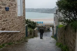 Mossy granite steps in a damp Dinard alley, local in rain jacket looking out toward misty beach cabins and Saint-Lunaire across the bay, early morning light highlighting wet textures and muted colors.