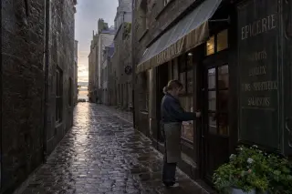 A narrow, rain-soaked street in Saint-Malo intra-muros at dawn, with a local woman unlocking an old grocery store under a weathered awning, soft golden morning light reflecting on wet cobblestones and granite walls.